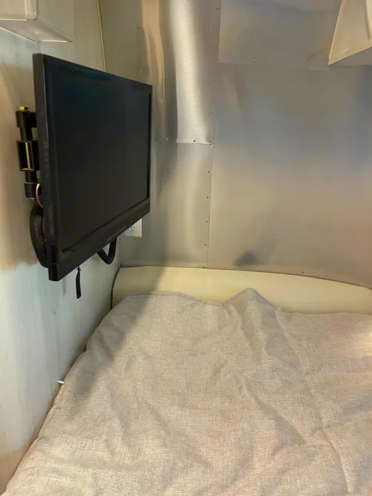 RV rental bedroom shows a wall-mounted black TV on aluminum walls, next to a bed with a light grey blanket.. Airstream Bambi 2022