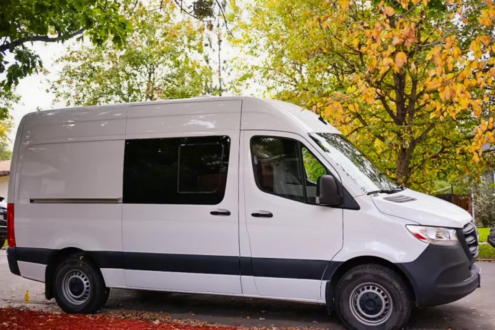 This white high-roof camper van, ideal for RV rental, is parked outdoors amidst trees with autumn leaves.. Mercedes-Benz Sprinter 2021