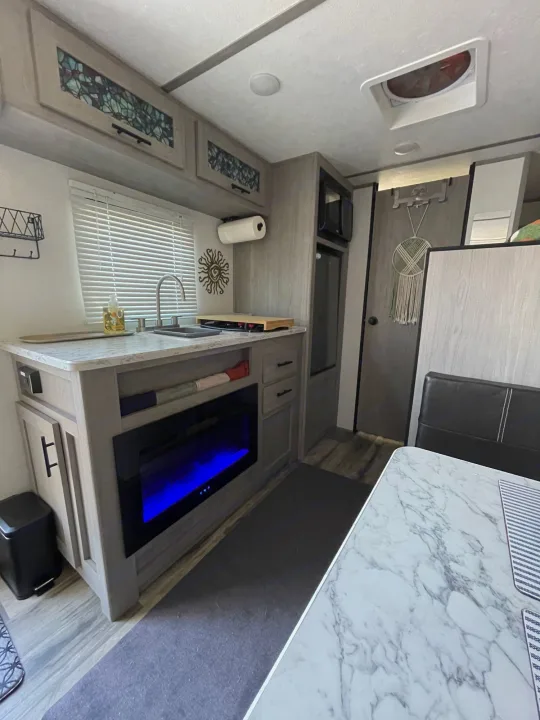 RV rental interior: kitchen with light cabinets, sink, blue fireplace, and dining area.. Keystone RV COLEMAN LANTERN LT 17B 2024