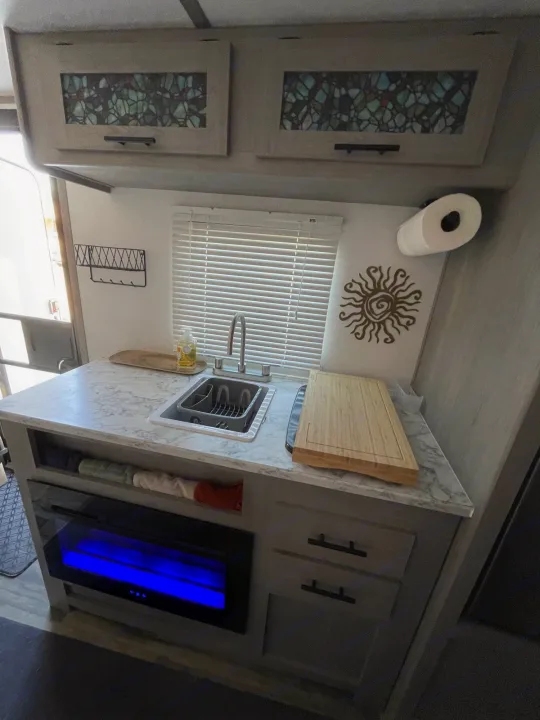 Inside an RV rental: a compact kitchen with a sink, decorative cabinets, cutting board, and blue-lit fireplace.. Keystone RV COLEMAN LANTERN LT 17B 2024