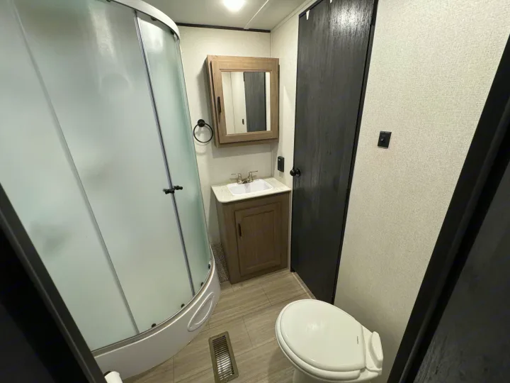 RV rental bathroom featuring a curved shower, vanity with sink, mirror, toilet, and modern dark wood door.. Keystone Hideout 2021