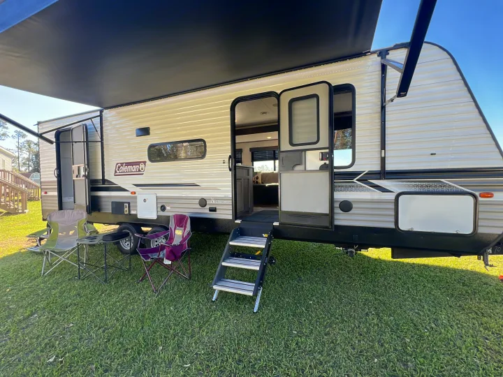 RV rental ready for adventure: A travel trailer with extended awning, open door, steps, and camp chairs on the grass.. Dutchmen Coleman Lantern 2023