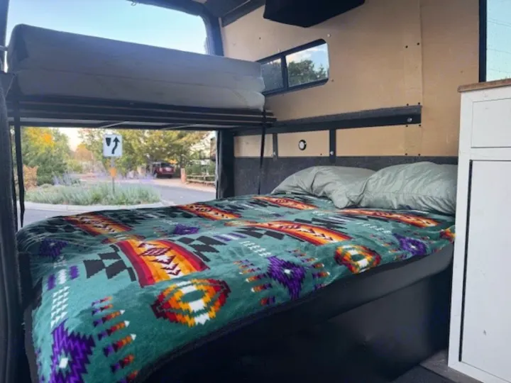 This RV rental's cozy interior features a vibrant blanketed bed below an upper bunk, with a clear view of the outdoors.. Mercedes-Benz Sprinter 2021