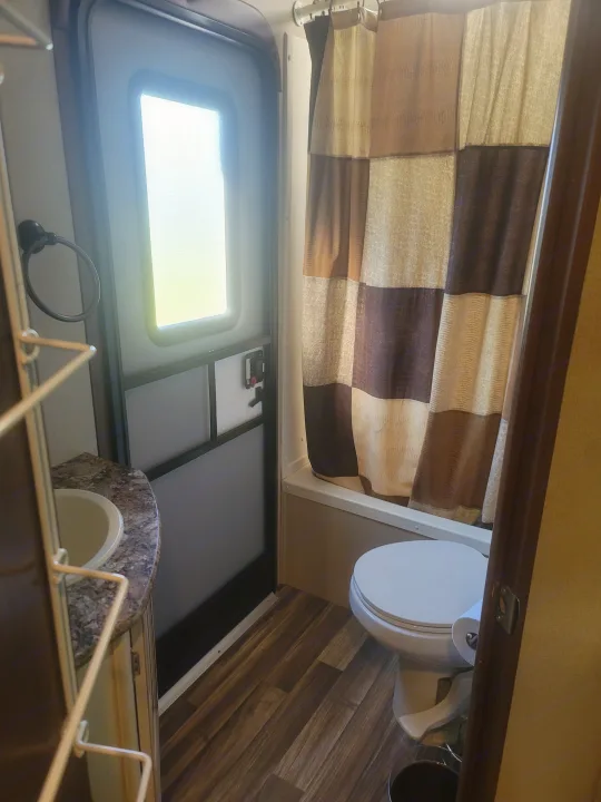 This clean RV rental bathroom features a compact sink, toilet, and shower with a brown curtain, lit by an exterior door.. Denali Lite 2015