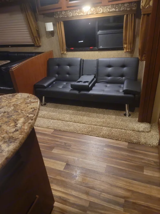 Inside this RV rental: a comfy living space with black futon (with cupholders), dining booth, wood floor, and a plush rug.. Denali Lite 2015