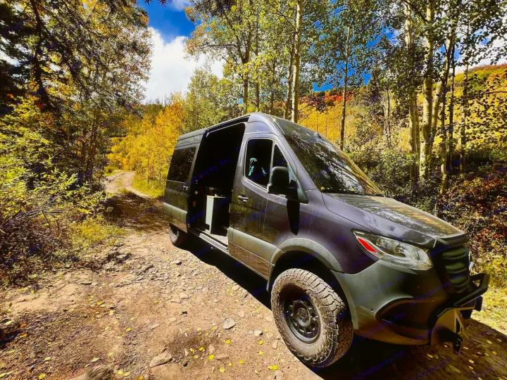 Fall adventure awaits! Dark rv rental van on a rugged dirt road surrounded by brilliant yellow autumn trees.. Mercedes Sprinter 2500 2024
