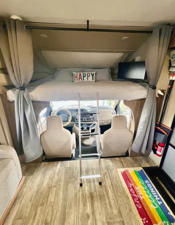 Cozy RV rental interior featuring an over-cab bunk bed, ladder, and bright front cabin view.. Jayco Envoy 100 2018
