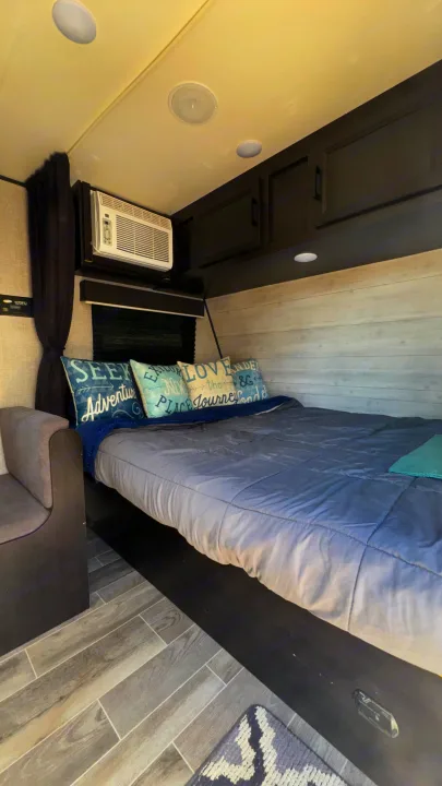 Cozy RV rental bedroom featuring a queen bed, decorative pillows, overhead cabinets, AC unit, and wood-look flooring.. Jayco Jay Flight SLX 2022