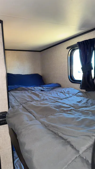 Interior of an RV rental: a cozy bunk bed with grey comforter, blue pillow, and a small window.. Jayco Jay Flight SLX 2022