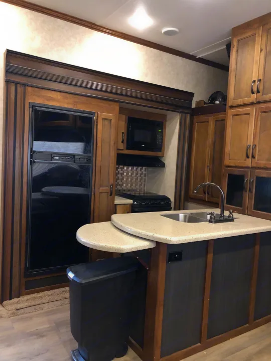 RV rental kitchen: modern interior with black appliances, light counters, island sink, and wood cabinets.. Keystone RV Carbon Toy Hauler 2015