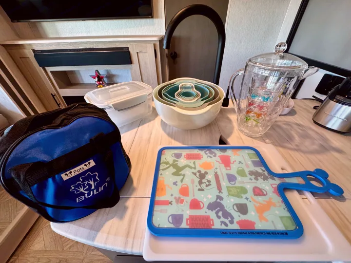 RV rental kitchen essentials: Bulin pots bag, nesting bowls, Disney cutting board, Mickey glasses, and storage containers.. Forest River Wildwood 2024
