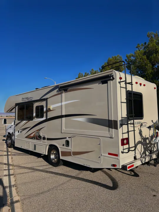 A light-colored Class C RV rental is parked on asphalt. The recreational vehicle has a ladder and a bike rack with bikes on the back.. Thor Motor Coach Freedom Elite 2017