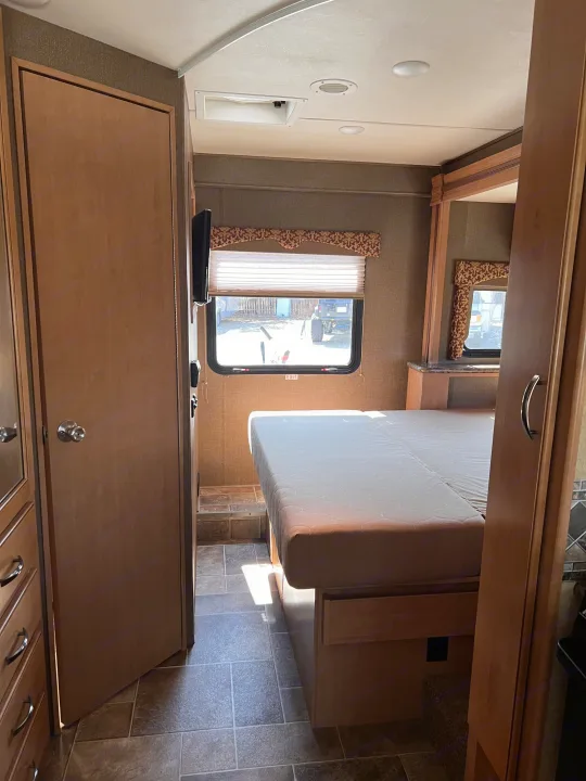 This RV rental's bedroom features a raised bed, wall-mounted TV, window with blinds, and light wood cabinetry.. Thor Motor Coach Freedom Elite 2017