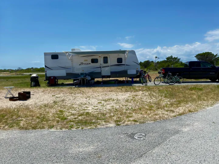 An RV rental setup at a sunny C6 coastal campsite with a travel trailer, truck, bikes, grill, and fire pit.. Crossroads RV Zinger 2016