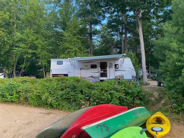 An inviting RV rental campsite features a white travel trailer, lush trees, and canoes & a kayak on a sandy path for outdoor adventures.. Crossroads RV Zinger 2016