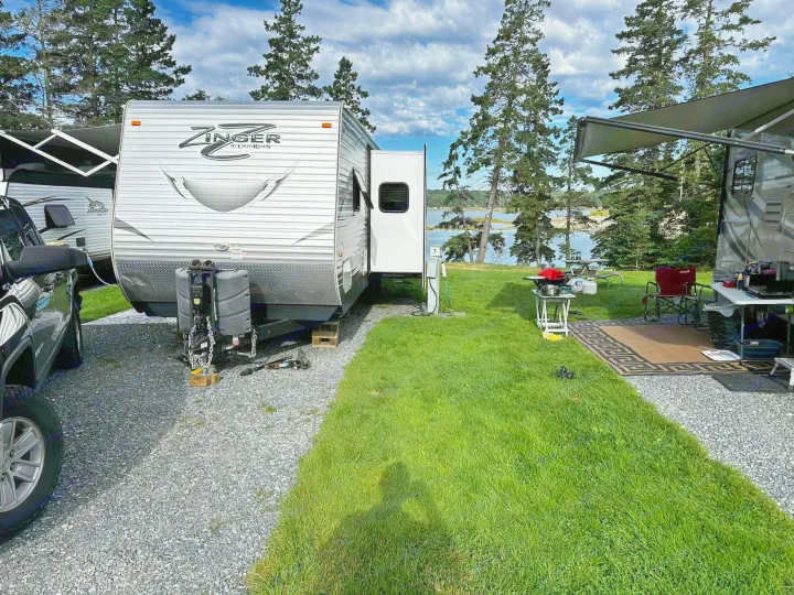 Spacious RV rental campsite with two travel trailers on gravel, leading to green grass, trees, and a water view.. Crossroads RV Zinger 2016