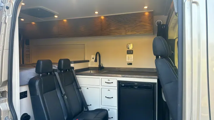Inside an RV rental: a compact living space featuring a kitchen, seating, bed area, and overhead storage.. RAM Promaster 2024