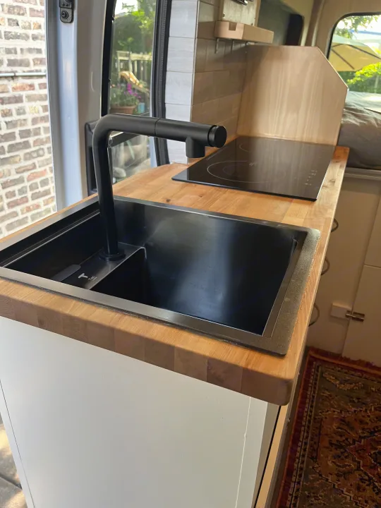 This RV rental van boasts a sleek kitchen: black sink, faucet, induction cooktop, and light wood counters.. Custom Built Transit 2017
