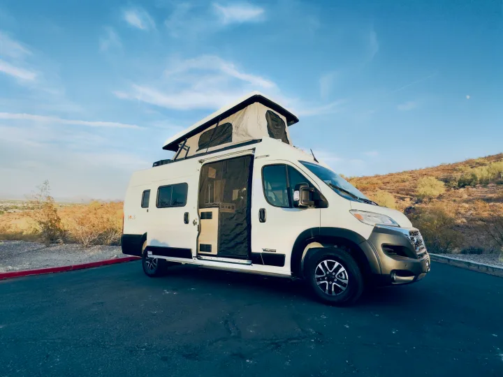White pop-top camper van for RV rental, with open side door, parked in sunny desert landscape.. Winnebago Solis 59PX 2025