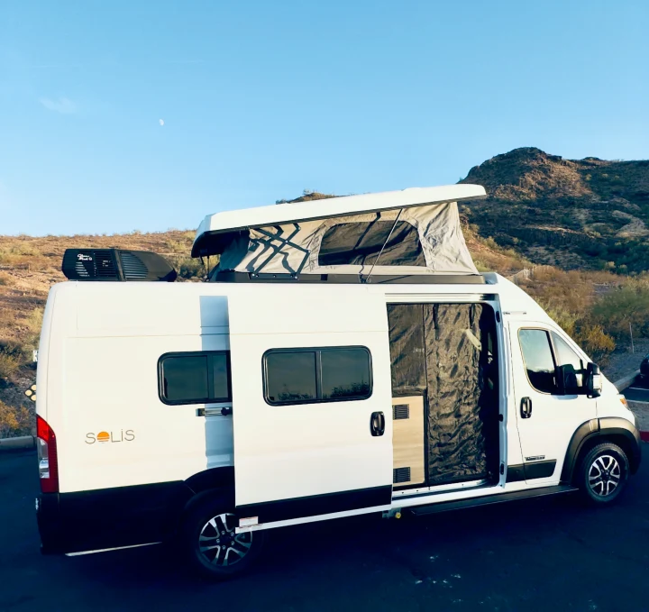 White Solis camper van with pop-top roof open, ready for your next RV rental. Parked by desert mountains under a blue sky.. Winnebago Solis 59PX 2025
