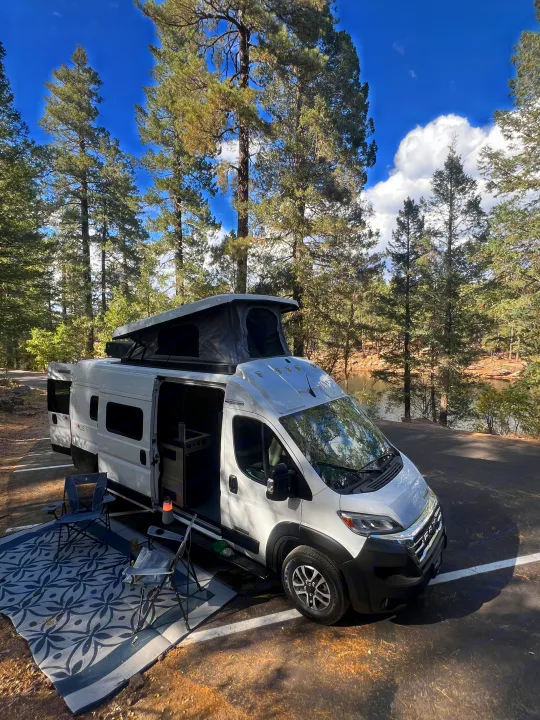 This RV rental camper van is perfectly set up for lakeside camping amidst tall pines, with chairs & an outdoor rug.. Winnebago Solis 59PX 2025