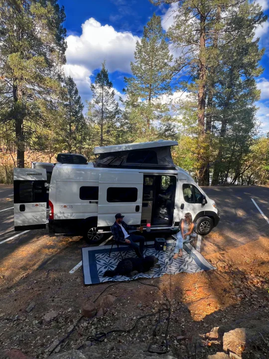 A couple and dog relax outside their white Winnebago Solis RV rental in a serene forest setting.. Winnebago Solis 59PX 2025