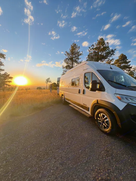 White RV rental on a gravel road at sunset, a person gazes at the golden field and distant view.. Winnebago Solis 59PX 2025