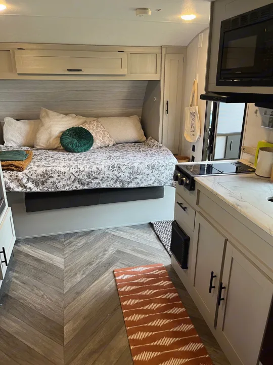 This cozy RV rental interior features a bed, compact kitchen area with stovetop, and wood-look flooring.. Forest River Wildwood FSX 2021