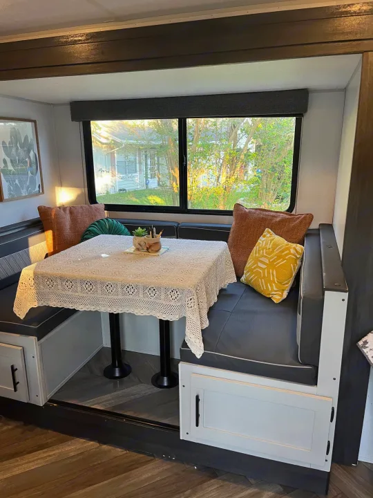 Cozy RV rental dining nook with gray benches, a lace-covered table, colorful pillows, and a bright window view.. Forest River Wildwood FSX 2021