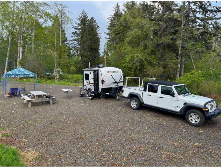 White Jeep Gladiator towing a travel trailer (RV rental) at a spacious gravel campsite with a blue canopy & picnic area.. Forest River Rockwood Geo Pro 2023