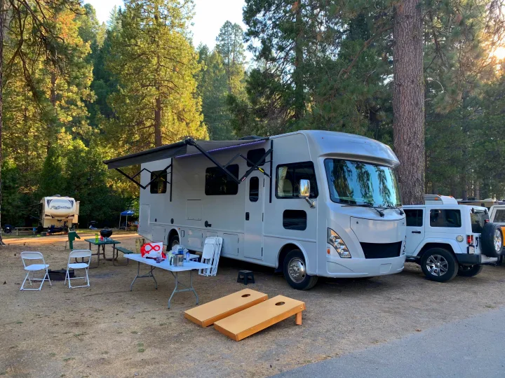 A white motorhome RV rental and Jeep parked at a sunny forest campground, with outdoor seating and cornhole games.. Thor A.C.E. 2018