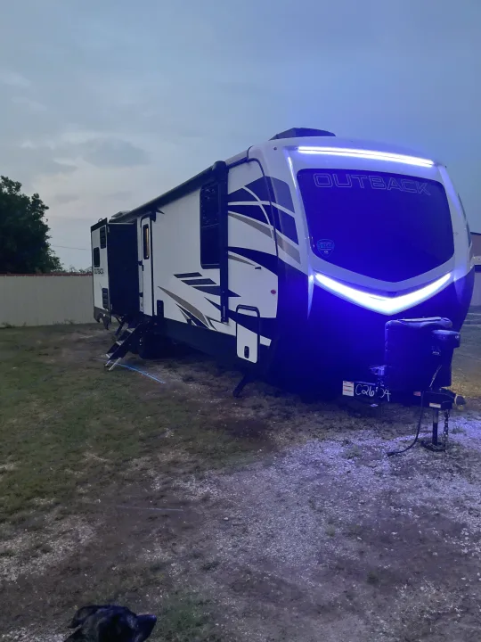 Night view of an RV rental: a modern Outback travel trailer with striking blue LED lights, parked on gravel and grass at dusk.. Keystone RV Outback Toy Hauler 2023