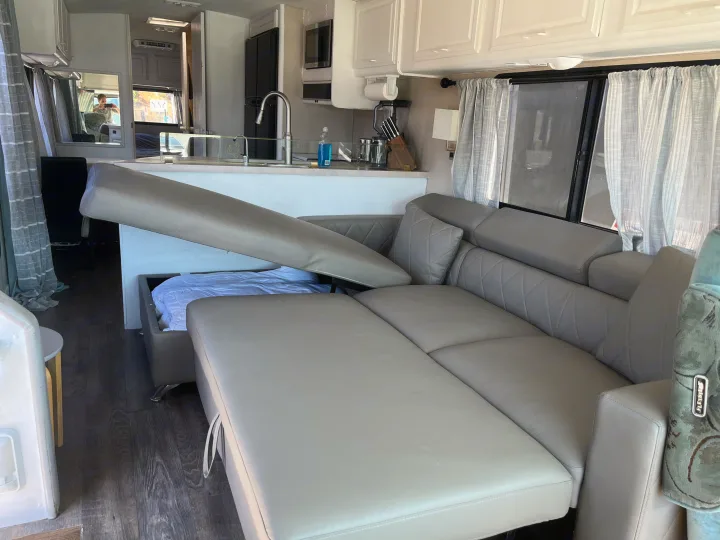 RV rental interior: A modern grey sofa bed is partially open, revealing a mattress and white bedding. A compact kitchen is visible beyond.. Fleetwood Bounder 1995