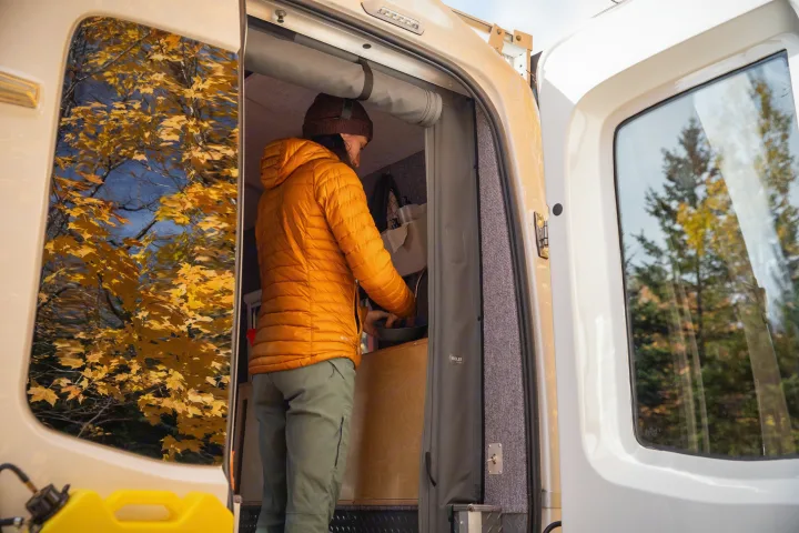 Inside an RV rental, a person in an orange jacket cooks. Bright fall foliage is seen through the open side door's window.. Gmc sierra 1500 2024
