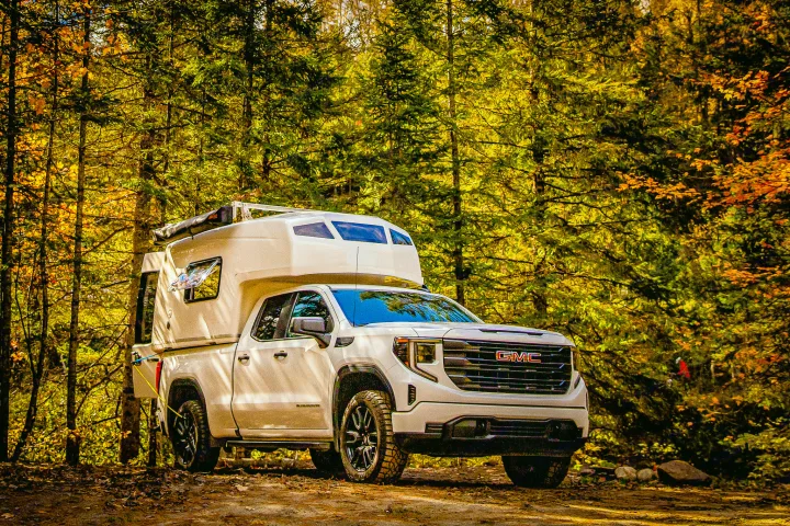 White GMC pickup with a truck camper on a dirt path in vibrant autumn woods. Consider this RV rental for your next adventure.. Gmc sierra 1500 2024