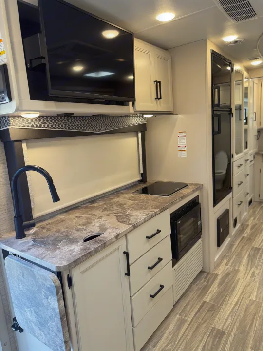 Modern rv rental interior: kitchen with sink, stovetop, microwave, fridge, TV, and light wood-look flooring.. Thor Vegas 2023