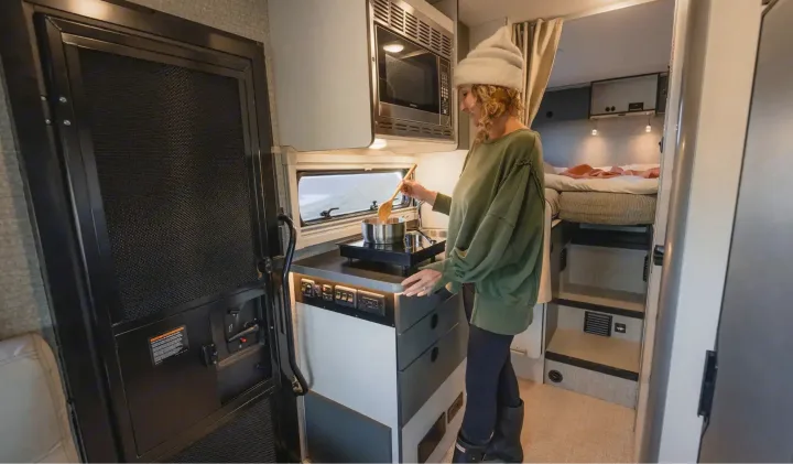 Inside an **rv rental**, a woman stirs a pot on a compact induction cooktop, highlighting functional mobile living.. Winnebago Ekko 2025