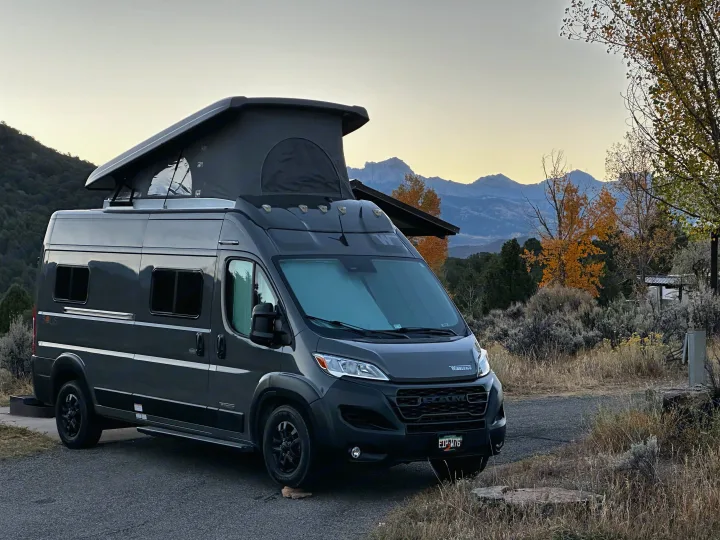 A dark gray pop-top camper van (RV rental) parked at a scenic campsite with mountains & autumn trees.. Winnebago Solis 2023