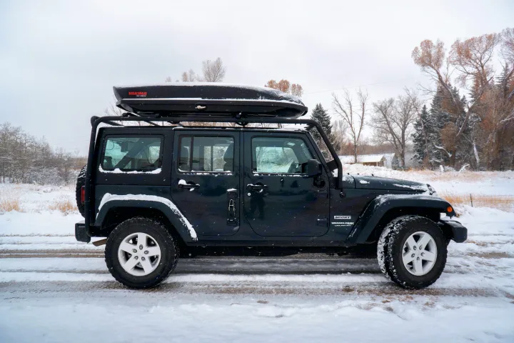 Dark Jeep Wrangler Unlimited with roof cargo box on a snow-covered road. Ideal winter adventure vehicle, perfect for an RV rental trip.. Jeep Wrangler Unlimited 2018