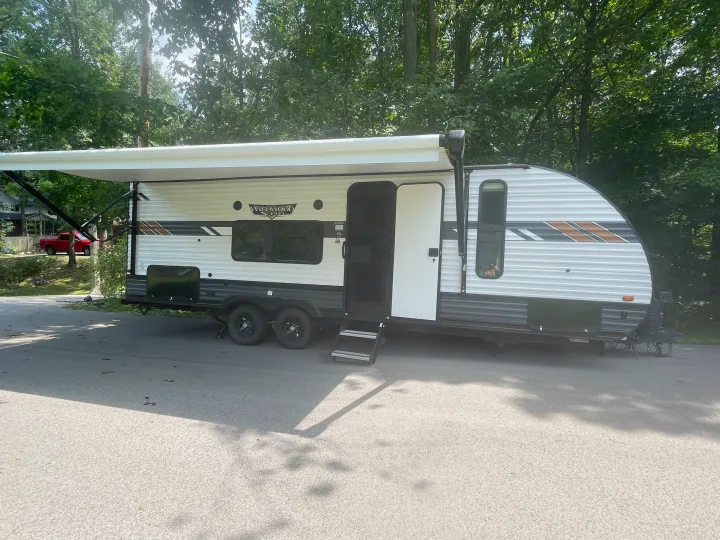 White Wildwood travel trailer RV rental with extended awning and open door, parked on asphalt in a treed setting.. Forest River Wildwood X-Lite 2022