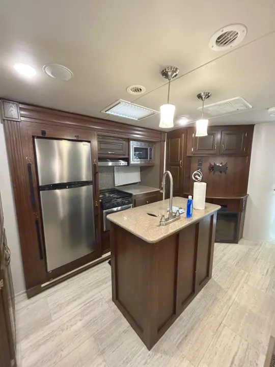 Modern rv rental kitchen with dark wood cabinets, stainless steel appliances, and island with sink.. Palomino Solaire 2018