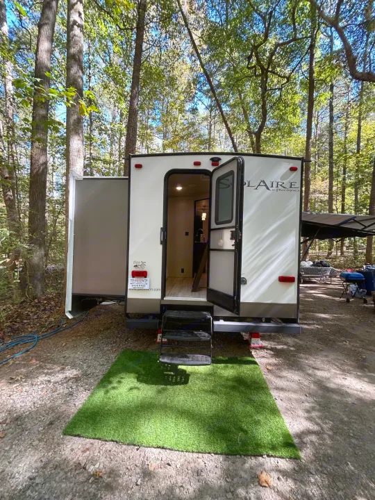 White travel trailer RV rental parked at a serene forest campsite with open door, showing interior and a green mat.. Palomino Solaire 2018