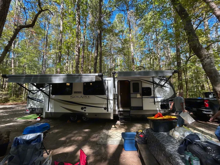 A white RV rental with extended awnings and slide-outs parked at a shaded, forested campsite with camping gear visible.. Palomino Solaire 2018