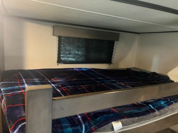 Cozy RV rental bunk bed featuring a plaid blanket, a window with a pulled-down blind, and a protective railing.. Grand Design Transcend 2021