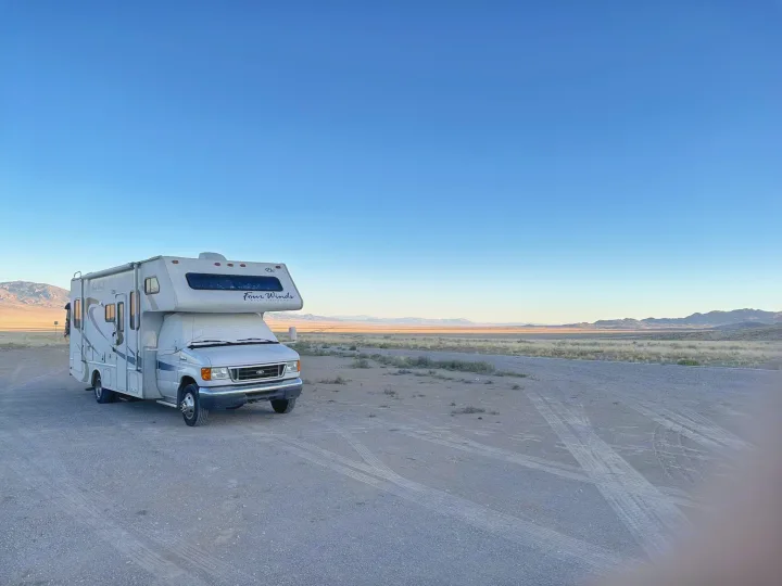 A white Class C rv rental motorhome is parked on a gravel road in a vast desert landscape with distant mountains.. Four Winds 5000 2008