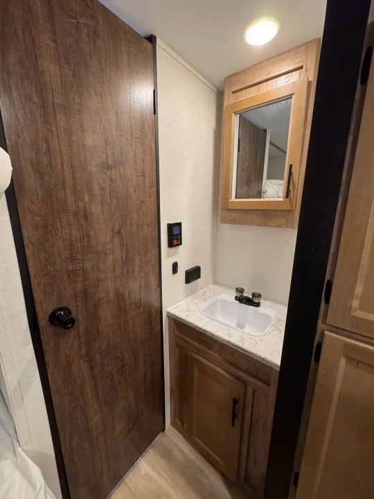 RV rental bathroom: features a compact sink, wood-toned vanity/mirror, a door, and wall controls.. Grand River 25BH 2025