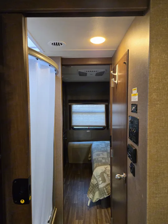 RV rental interior showing a compact hallway leading to a cozy bedroom, with a shower on the left and control panels on the right.. Thor A.C.E. 2018