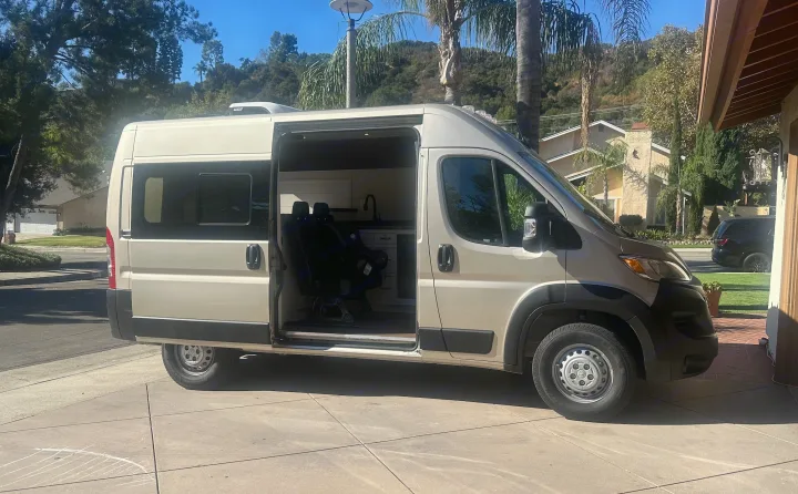 A modern beige camper van for rv rental, parked with its side door open, revealing a custom interior with seating and a kitchen.. RAM Promaster 2024