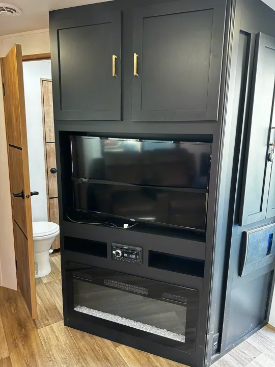 RV rental's sleek black entertainment unit: cabinets, TV, stereo, and electric fireplace, next to a small bathroom.. Keystone RV Outback Toy Hauler 2023