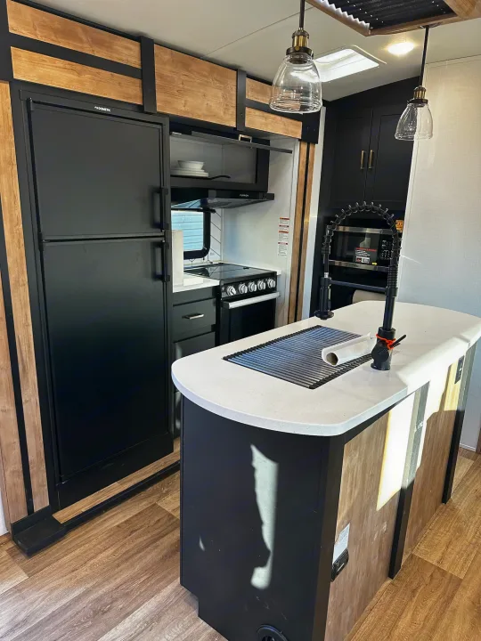 Modern RV rental kitchen with black Dometic fridge, stove, island with sink, and wood-look cabinetry.. Keystone RV Outback Toy Hauler 2023